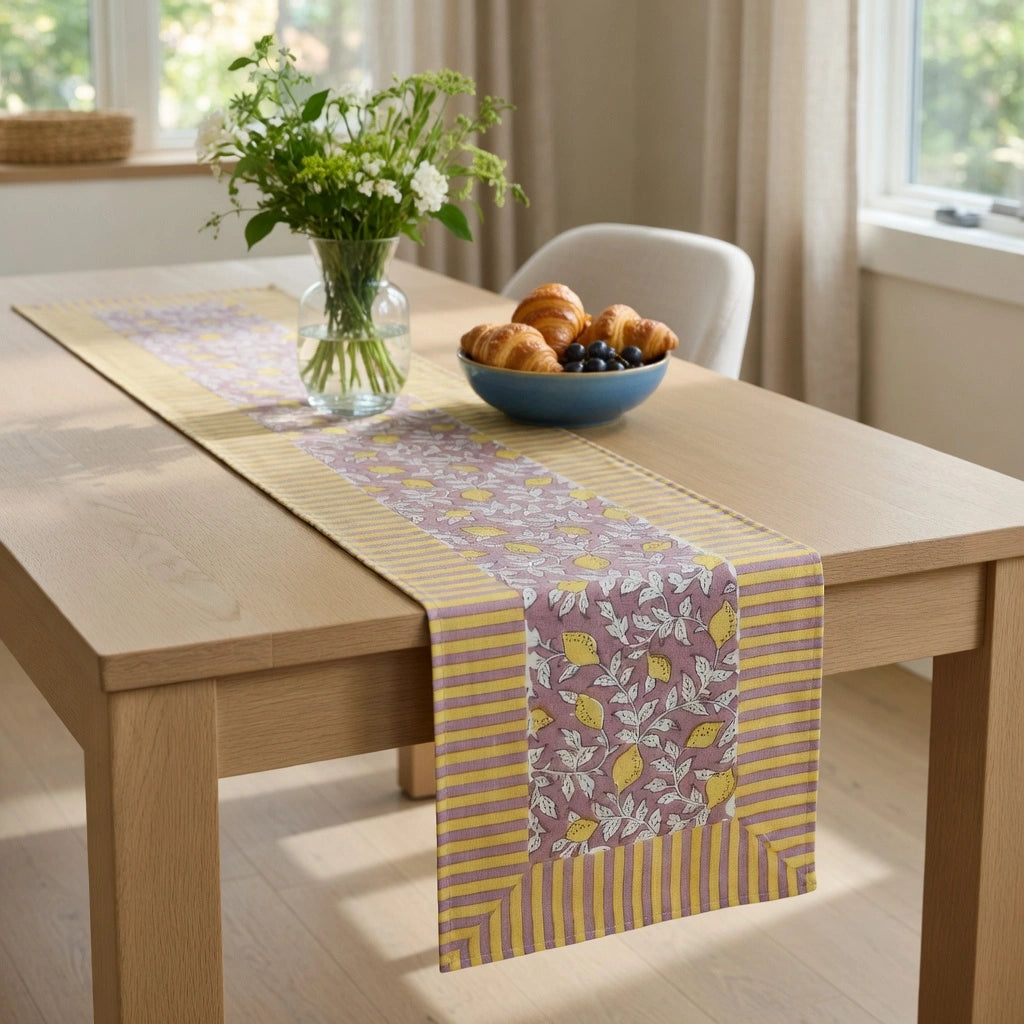 A rectangular table runner with a yellow and lavender floral pattern and striped borders, styled on a light wood dining table with a vase of flowers and a bowl of pastries.