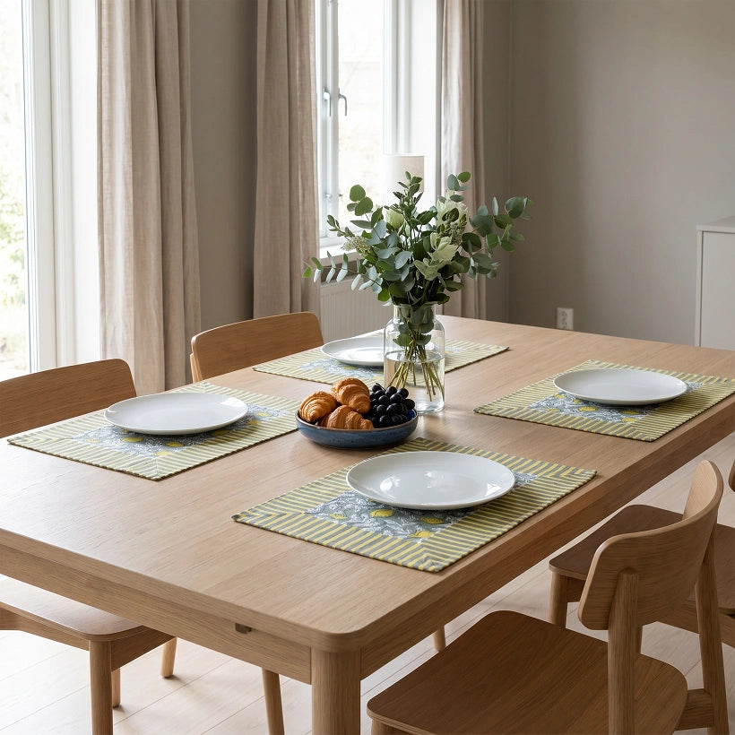 A light oak wood dining table set with four green patterned placemats, white plates, a bowl of fruit, and a vase of greenery, shown in a bright, modern dining room.