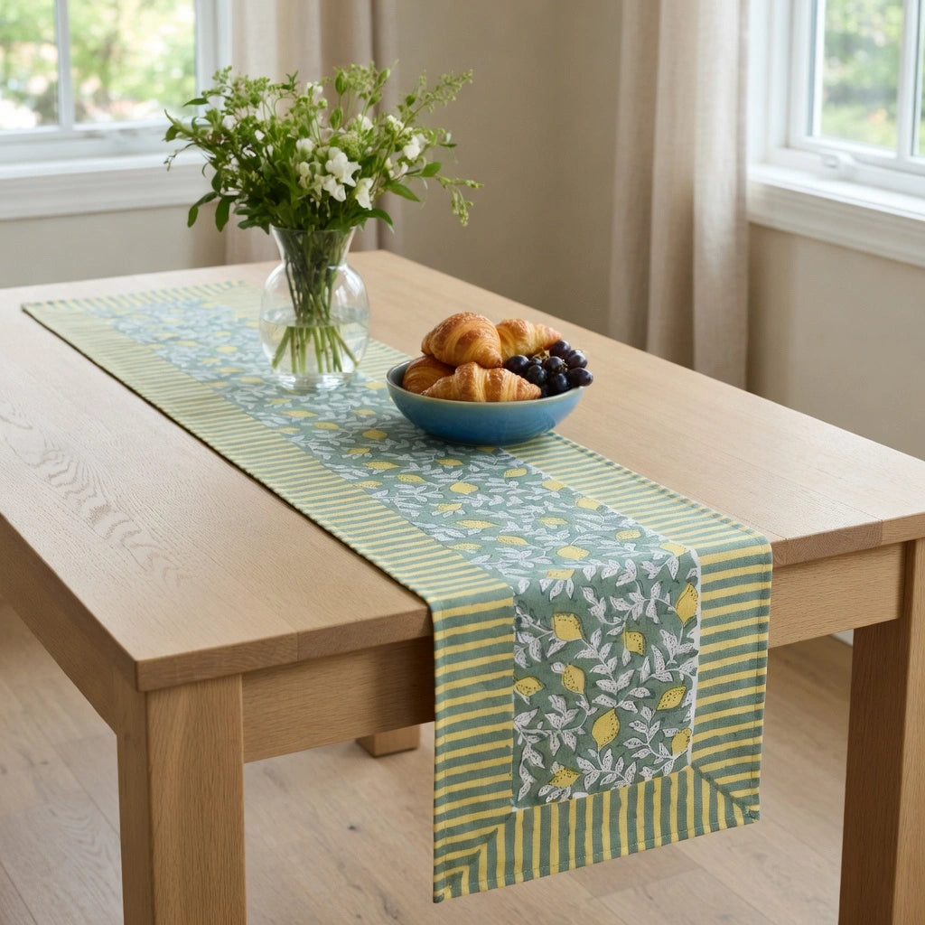 A rectangular table runner with a green and yellow floral and striped pattern is draped over a light wood dining table. The runner features a central panel with yellow lemons and white flowers, bordered by green and yellow stripes.