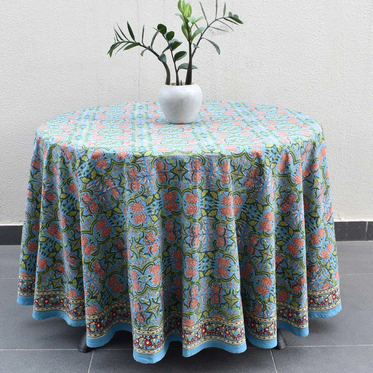 A round table covered with a light blue tablecloth featuring a colorful floral and ornamental pattern, styled with a white vase containing green leaves, set against a neutral wall and tiled floor.