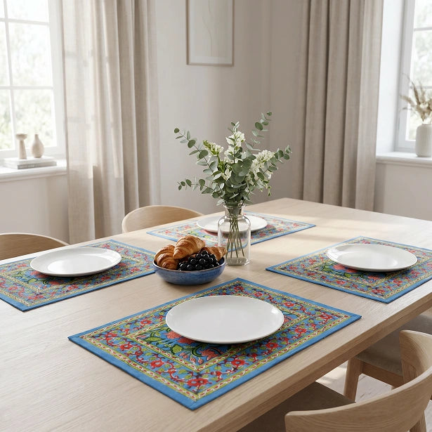 A dining table set with four colorful floral placemats, white plates, a glass vase of flowers, and a bowl of pastries in a bright, modern dining room.