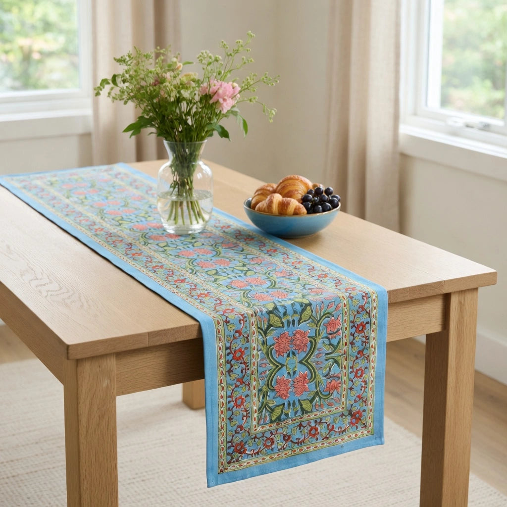 A rectangular table runner with a light blue border and intricate floral patterns in pink, green, and gold, styled on a light wood dining table in a bright room.