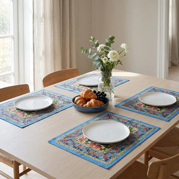 Four colorful floral table placemats with white plates, arranged on a light wood dining table set for a meal, with a vase of white flowers and croissants in the center.