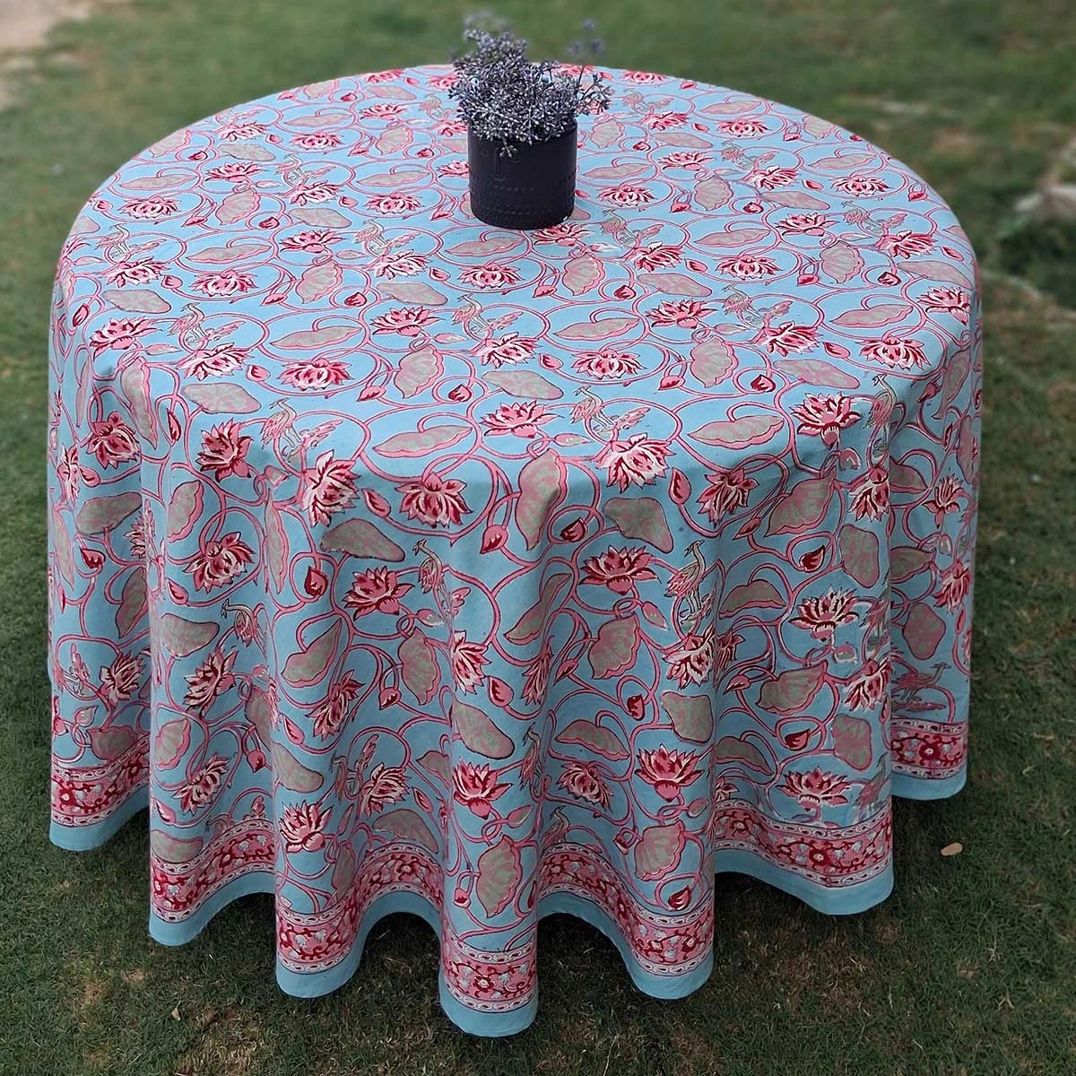 A round table covered with a light blue tablecloth featuring a red and pink floral pattern, set outdoors on green grass with a small black pot on top.