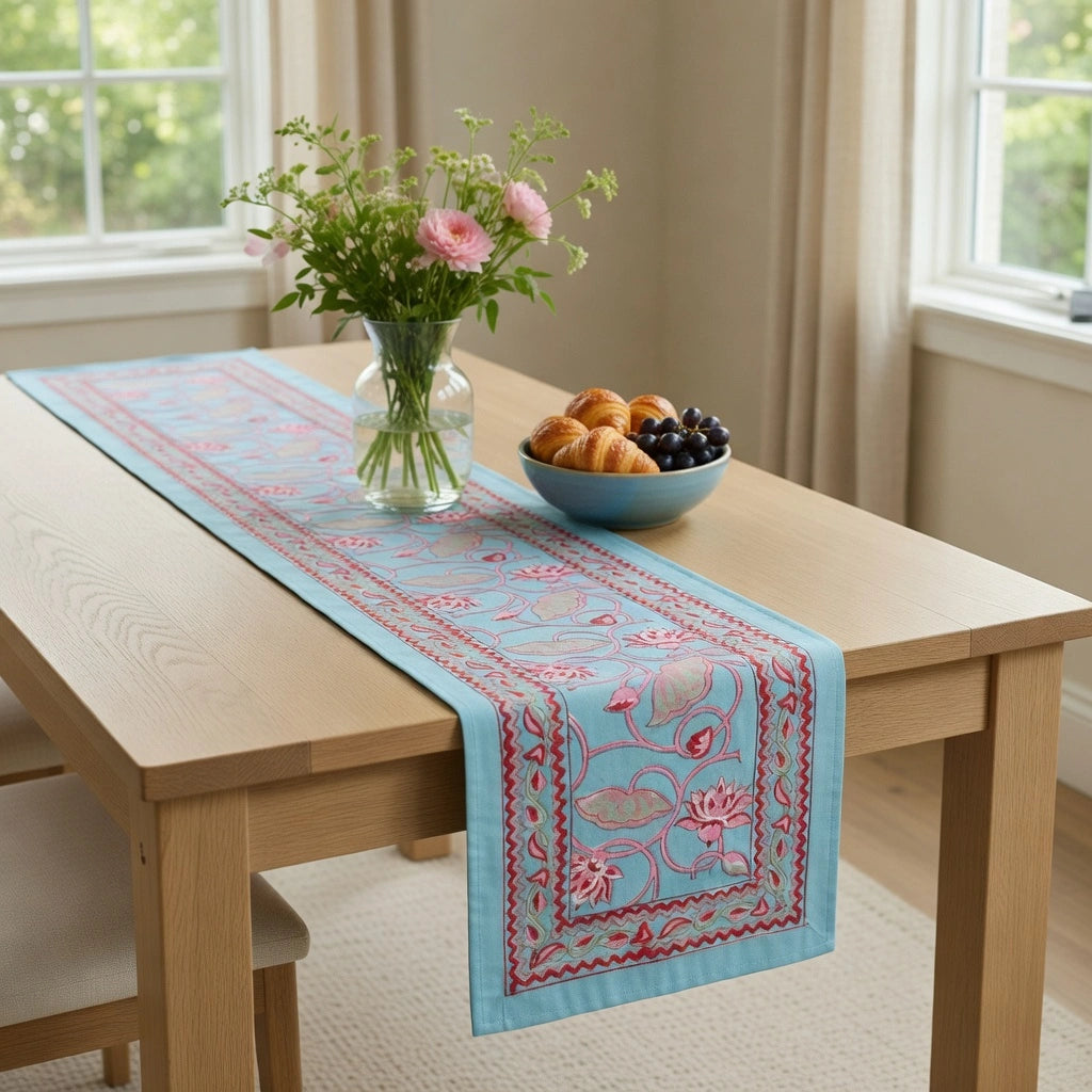 A light blue embroidered table runner with pink floral patterns and red borders, styled on a light wood dining table with a vase of flowers and a bowl of fruit in a bright room.
