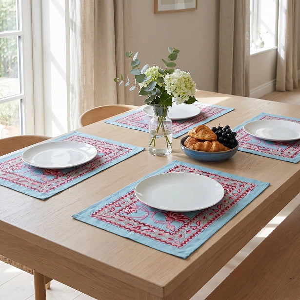 A dining table set with four light blue placemats featuring red embroidered patterns, white plates, a glass vase with white flowers, and a bowl of pastries in a bright, sunlit room.