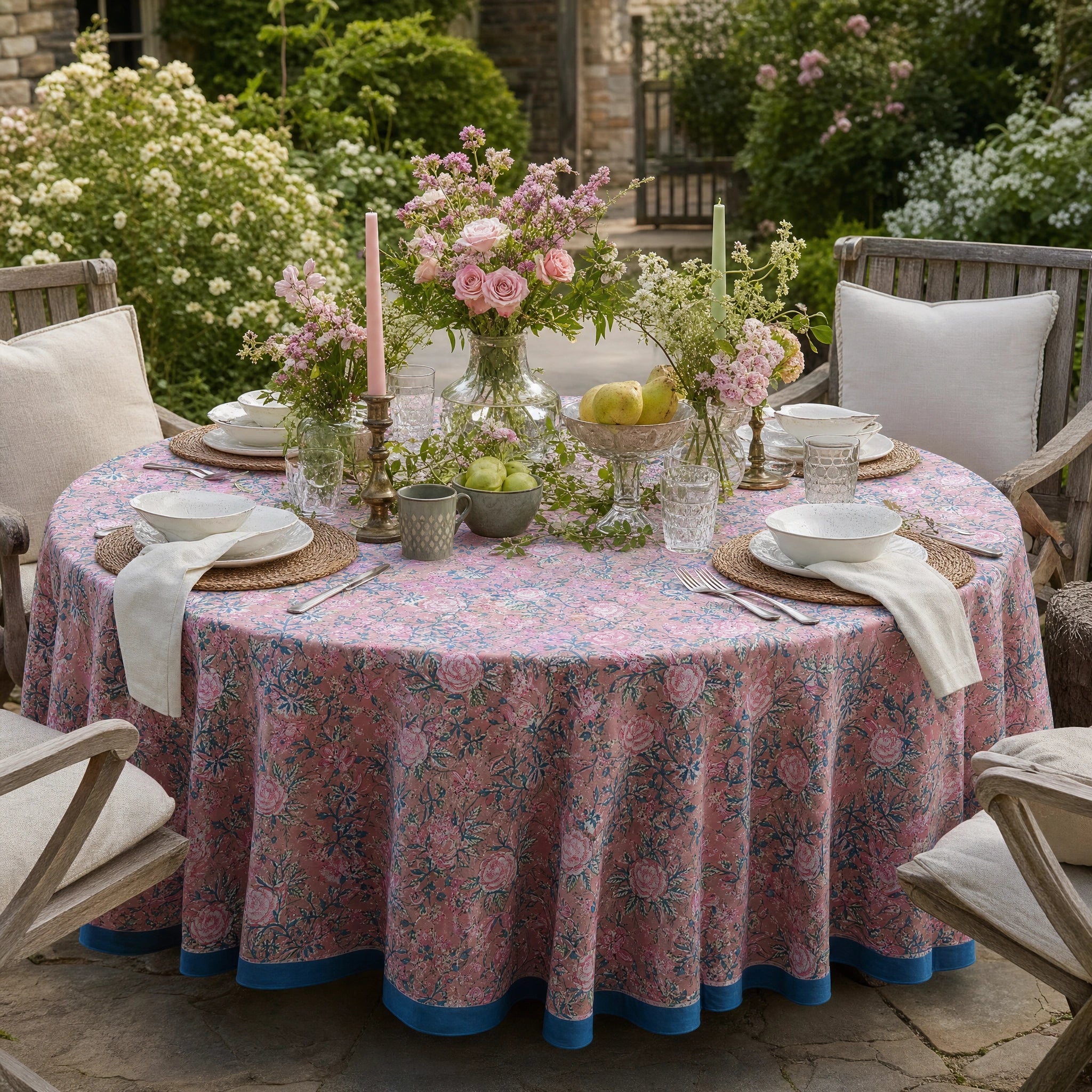 A round outdoor dining table set with a pink floral tablecloth, white plates, folded napkins, woven placemats, glassware, and floral centerpieces, surrounded by wooden chairs in a garden setting.