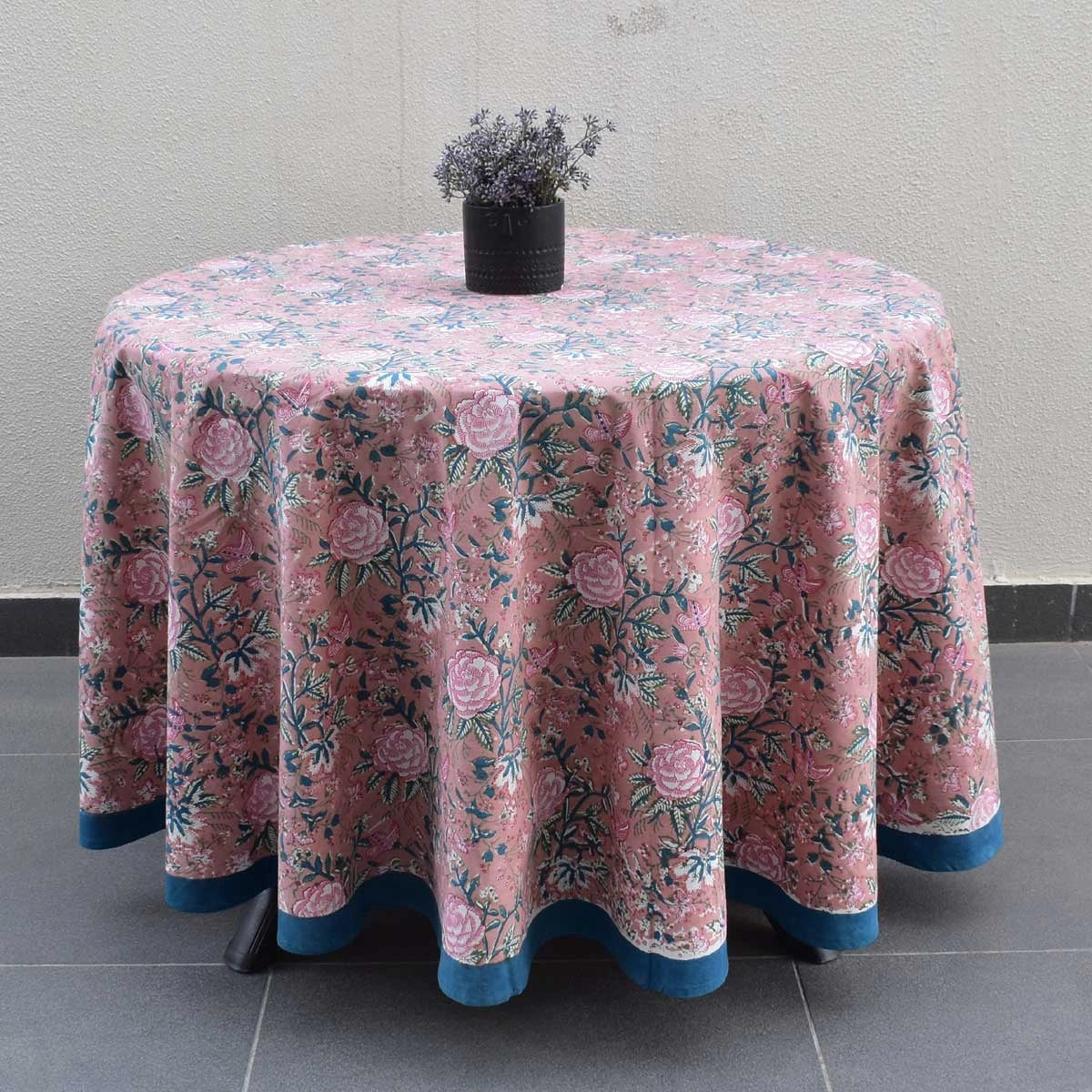 A round table covered with a pink floral tablecloth featuring blue and green botanical patterns and a blue ruffled edge, with a small black vase of flowers on top, set against a neutral wall and tiled floor.