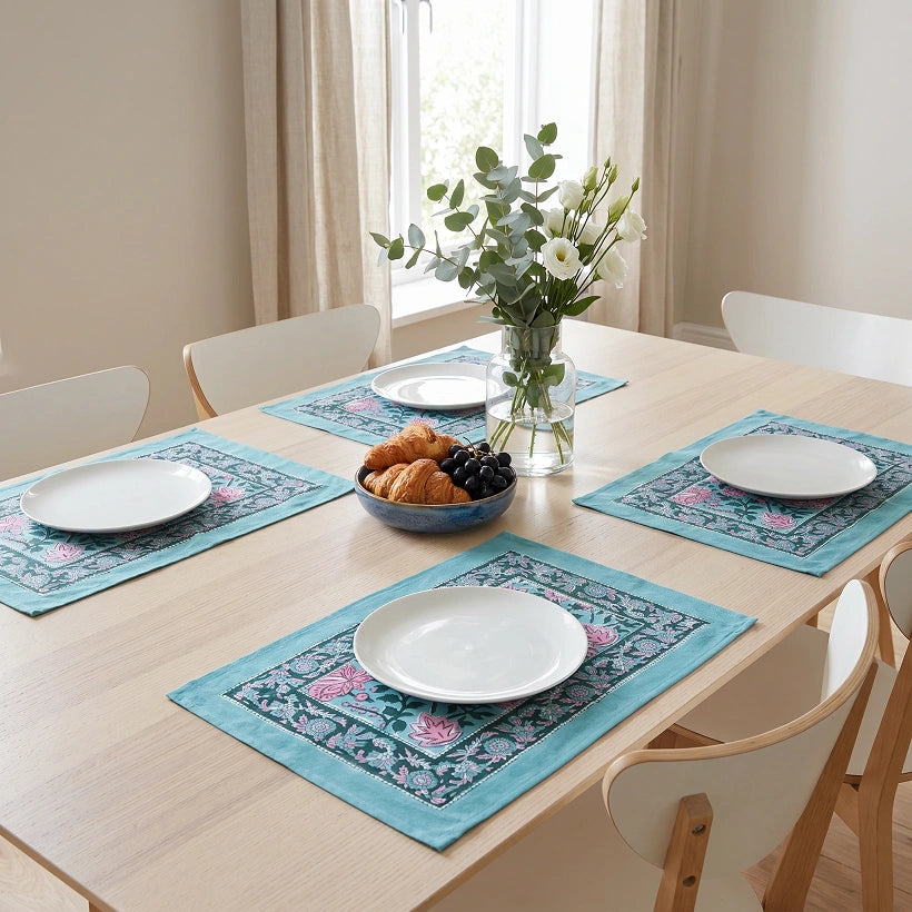 Four matching rectangular table placemats with a turquoise blue border and pink floral pattern, arranged on a light wood dining table set with white plates, a bowl of pastries and grapes, and a vase of flowers.