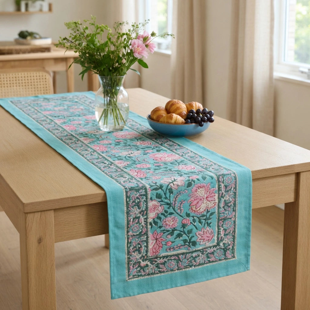 A turquoise table runner with pink floral embroidery and a detailed border, styled on a light wood dining table with a vase of flowers and a bowl of fruit in a bright, neutral room.