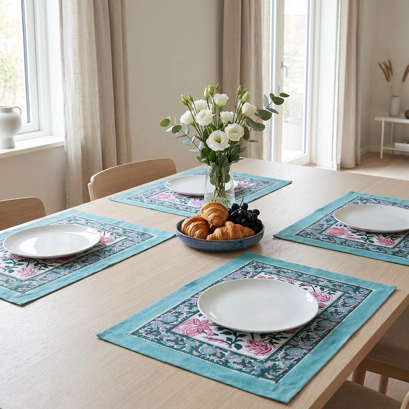 A dining table set with four matching turquoise floral placemats, white plates, a bowl of pastries and grapes, and a vase of white flowers in a bright, modern dining room.