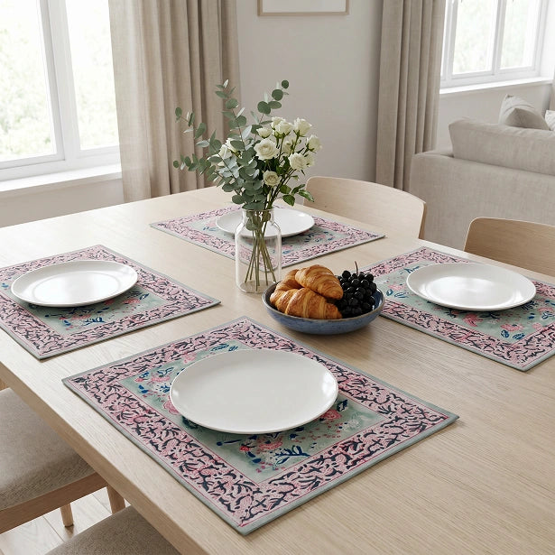 Four rectangular table placemats with a pink, blue, and beige floral border pattern are arranged on a light wood dining table, each styled with a white plate, a glass vase of flowers, and a bowl of pastries in a bright dining room.
