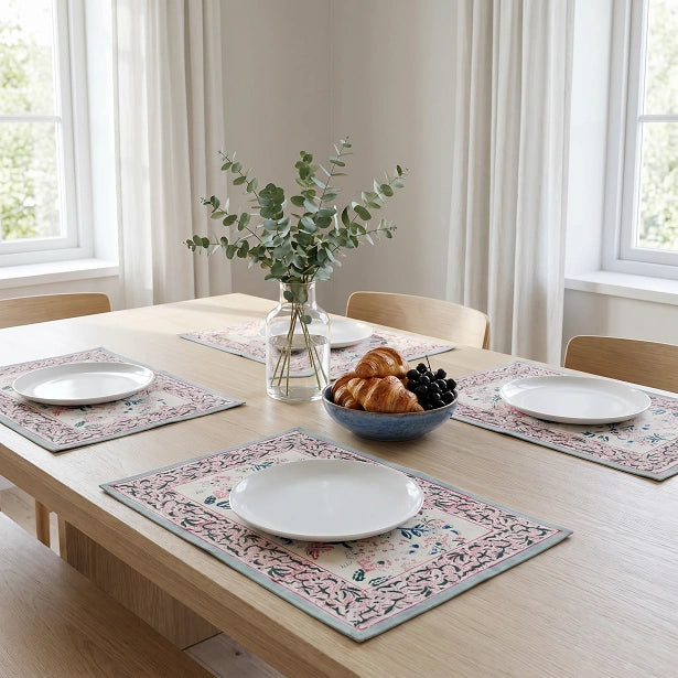 Four rectangular floral placemats with white plates are arranged on a light wood dining table in a bright room. A glass vase with eucalyptus stems and a blue bowl of croissants and black grapes sit in the center.
