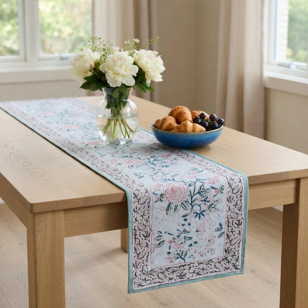 A rectangular table runner with a delicate floral and botanical print in soft blue, pink, and green tones, styled on a light wood dining table. The runner features a central floral panel and an ornate border, shown in a bright, modern dining room setting alongside a vase of white flowers and a blue bowl of pastries.