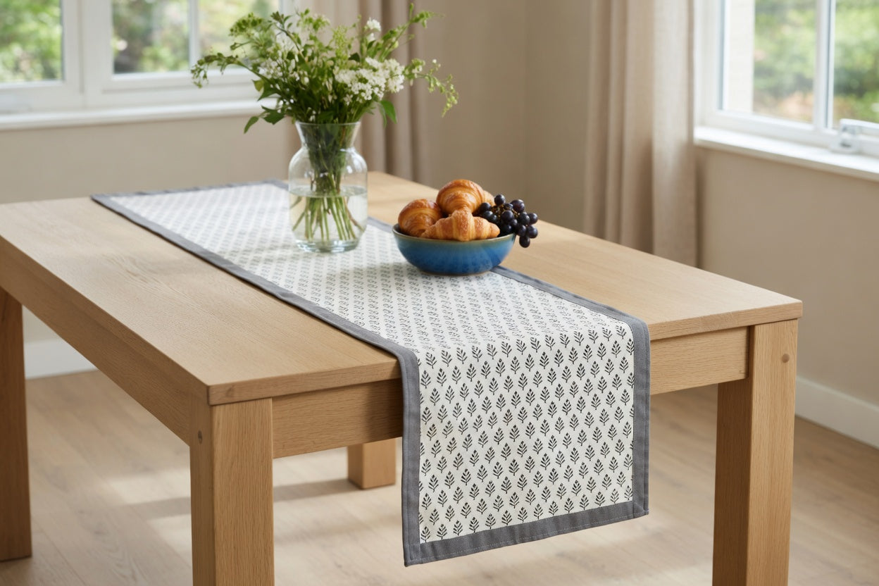 A light wood dining table in a bright room, styled with a white table runner featuring a gray geometric pattern and a gray border. A glass vase with white flowers and a blue bowl of fruit sit on the table.