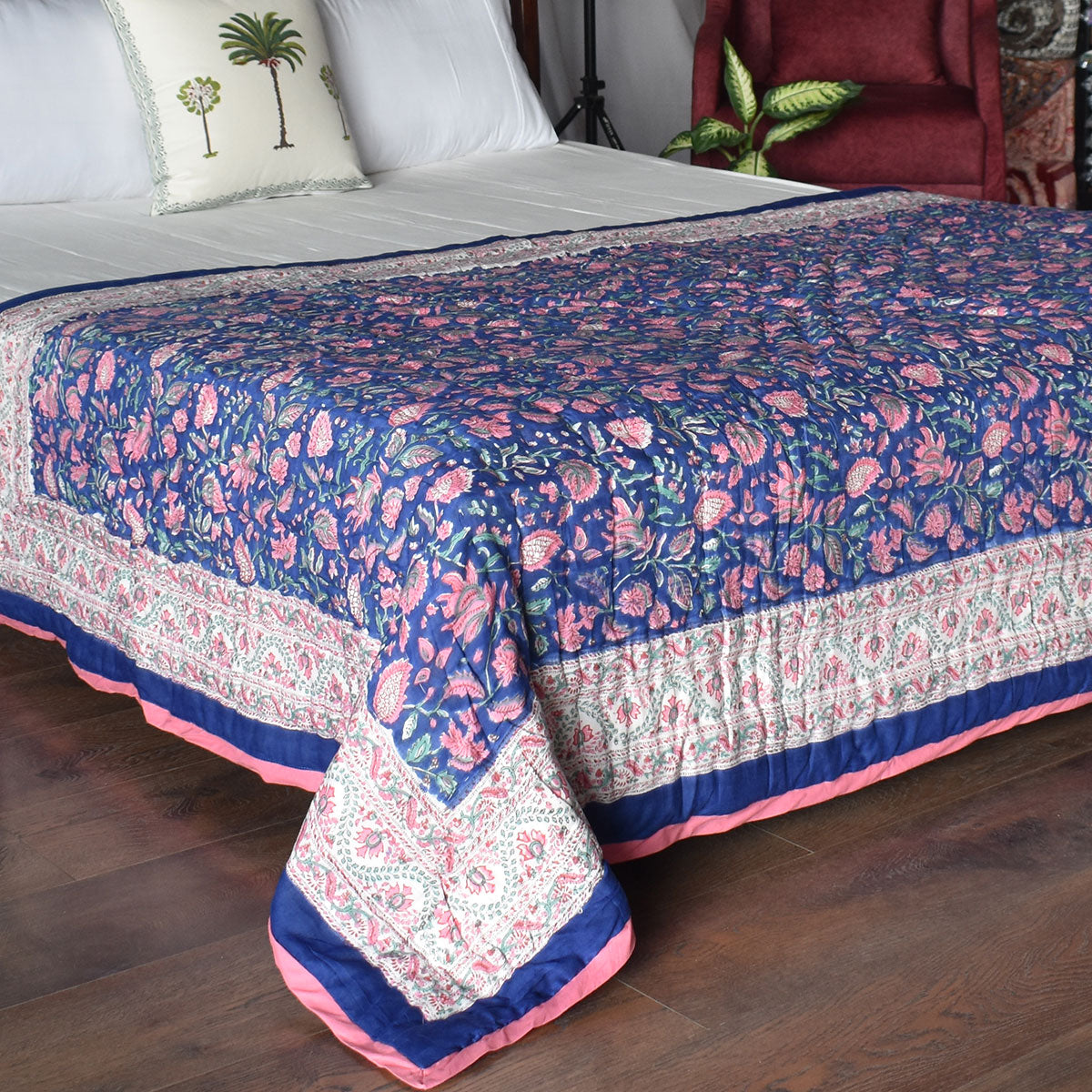 A blue quilted bedspread with pink and white floral and paisley patterns, styled on a bed in a bedroom setting with wooden floors, a red armchair, and decorative pillows.