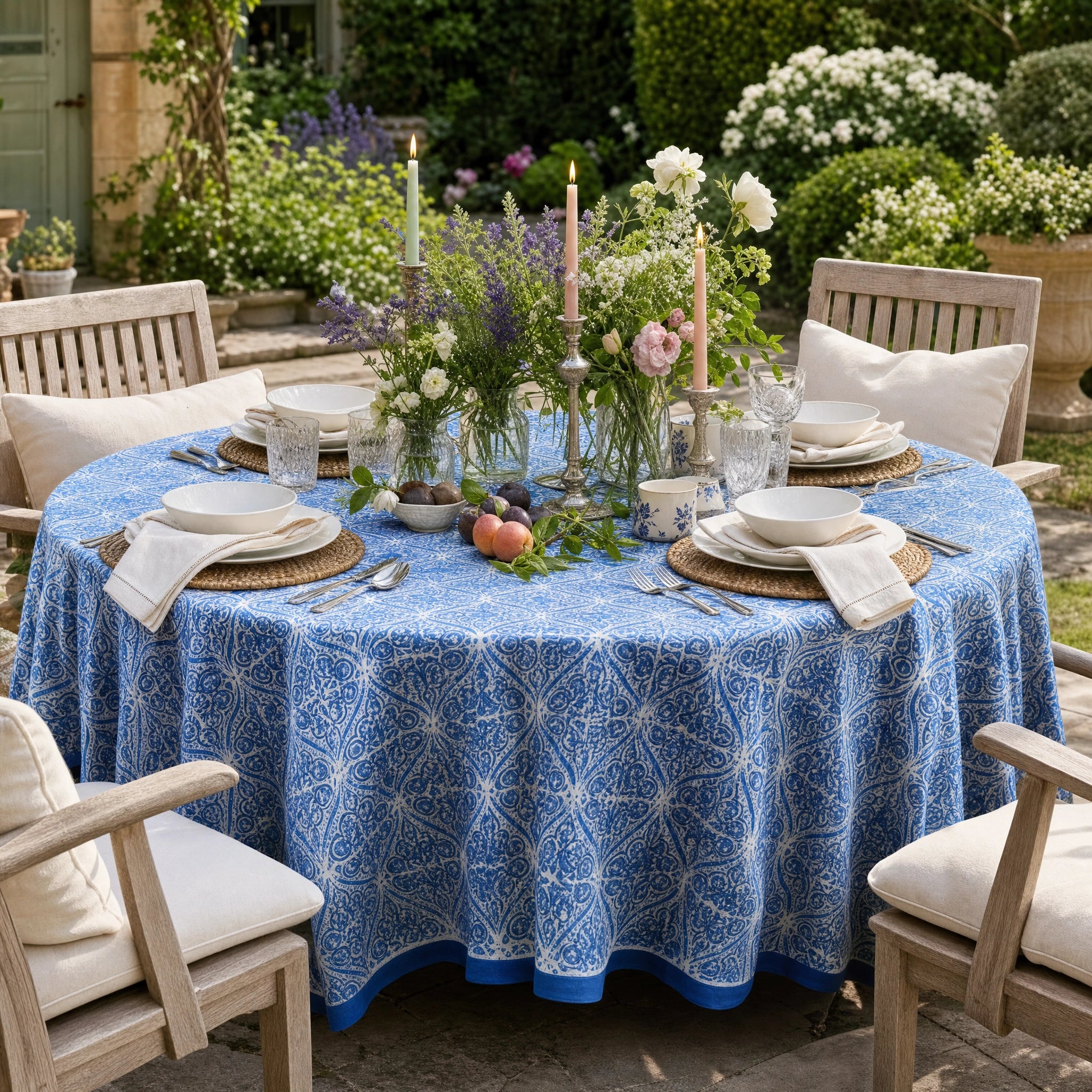 A round outdoor dining table covered with a blue tablecloth featuring a white geometric floral pattern, set with white plates, folded napkins, woven placemats, and a floral centerpiece in a garden patio setting.
