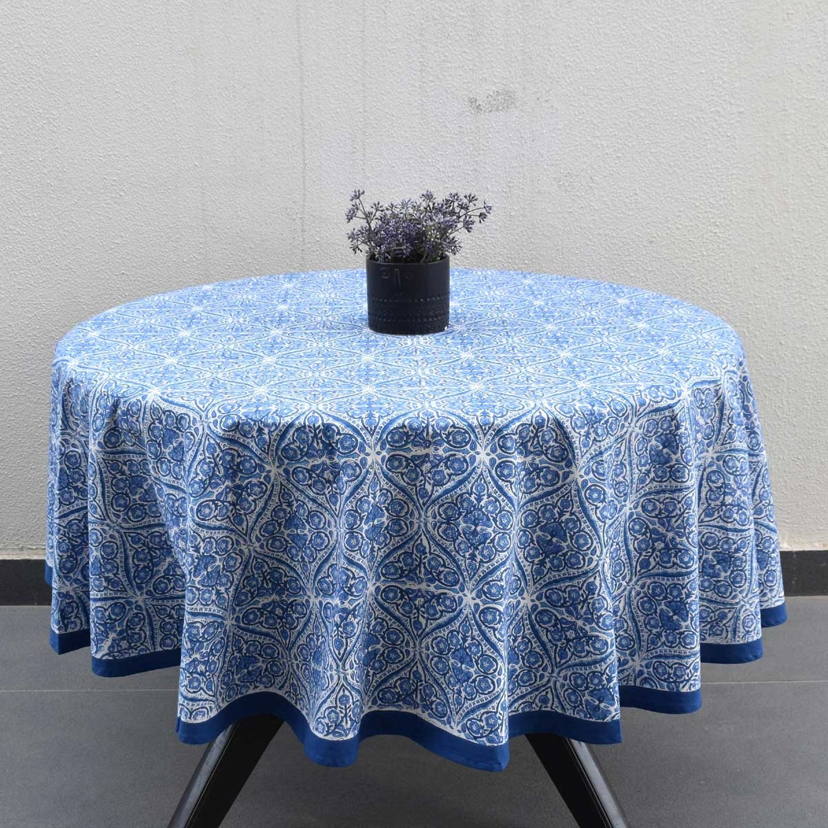 A round table covered with a blue and white patterned tablecloth, with a small black planter holding dried lavender placed on top, set against a light gray textured wall.