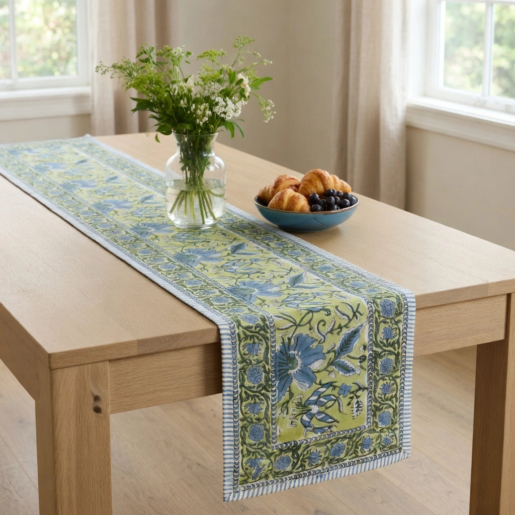 A rectangular table runner with a blue floral and vine pattern on a yellow-green background, styled on a light wood dining table with a vase of flowers and a bowl of pastries.