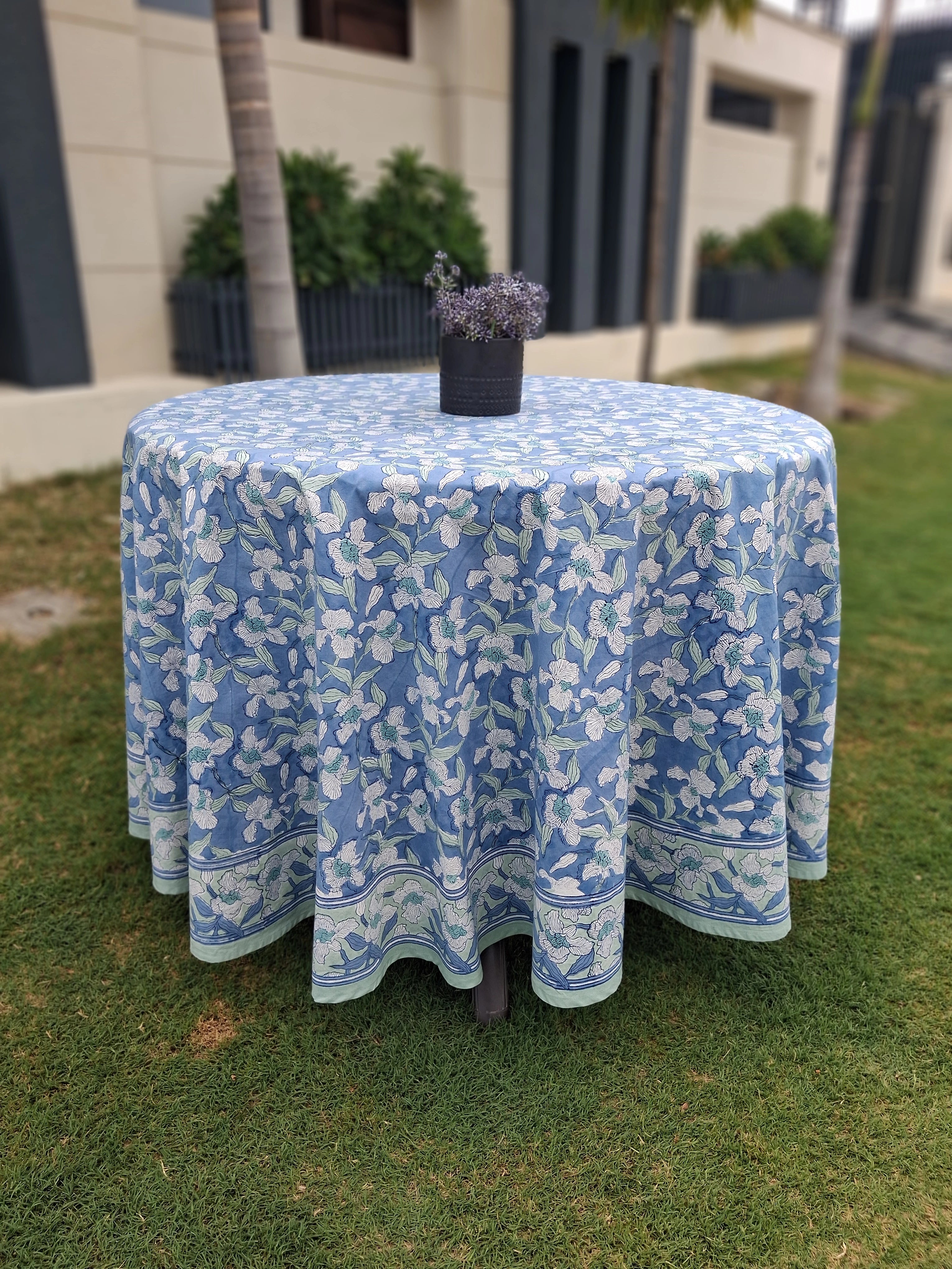 A round table draped in a light blue tablecloth with a white floral pattern, set outdoors on a green lawn with a small potted plant on top.