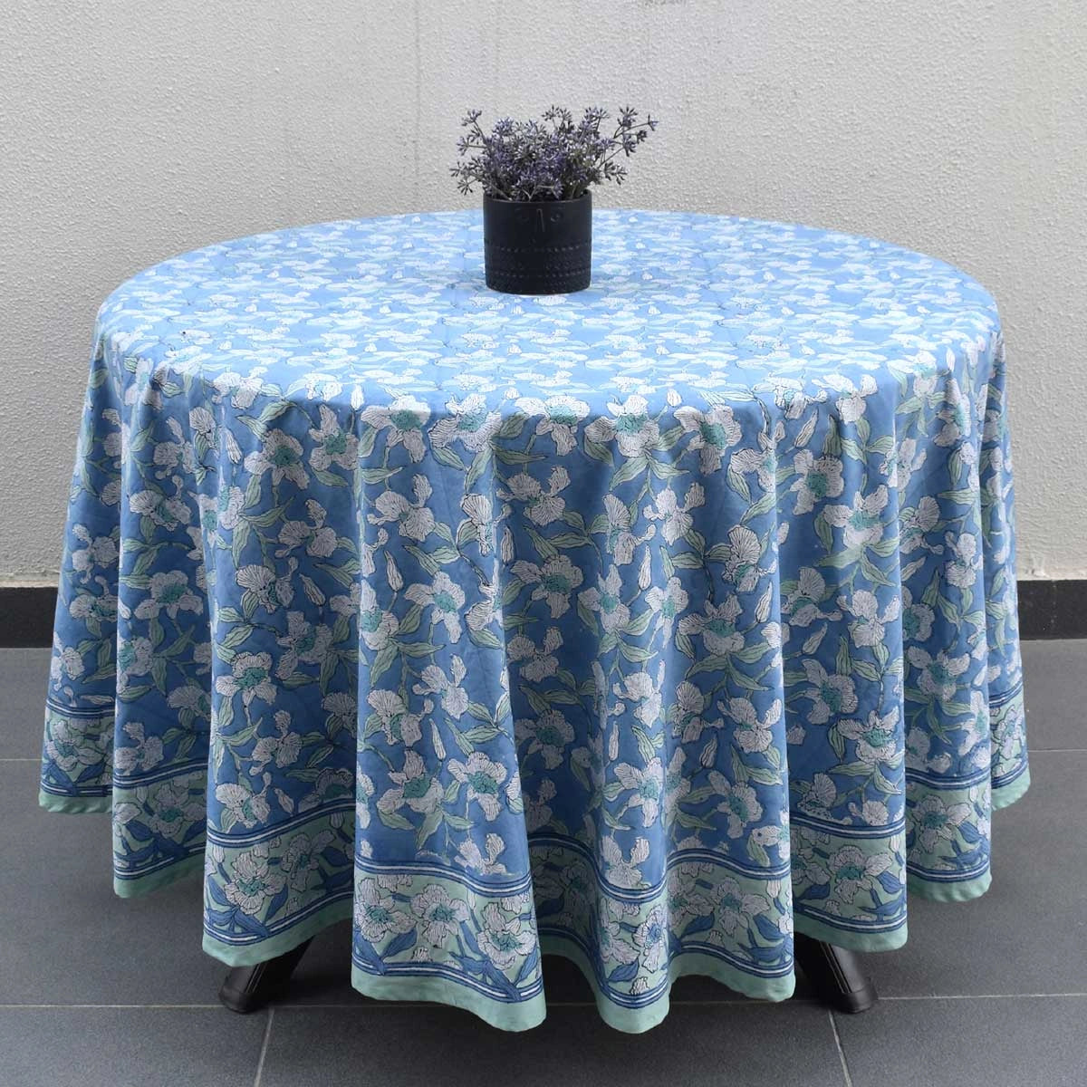 A round table covered with a light blue floral tablecloth, styled with a small black pot containing dried lavender, set against a neutral gray wall and tiled floor.