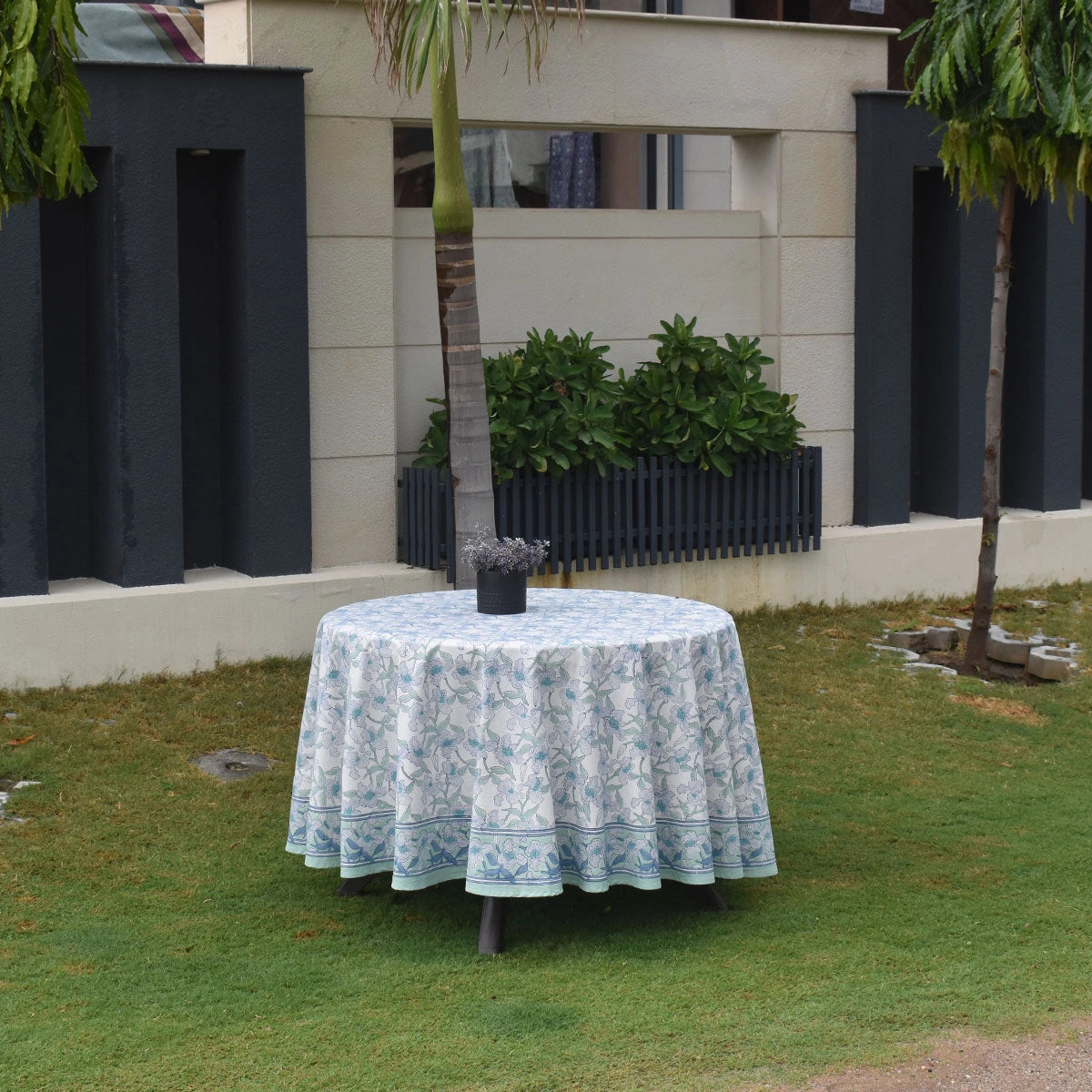 A round outdoor table covered with a light blue and white floral patterned tablecloth, set on a green lawn in front of a modern building with dark gray columns and a palm tree.