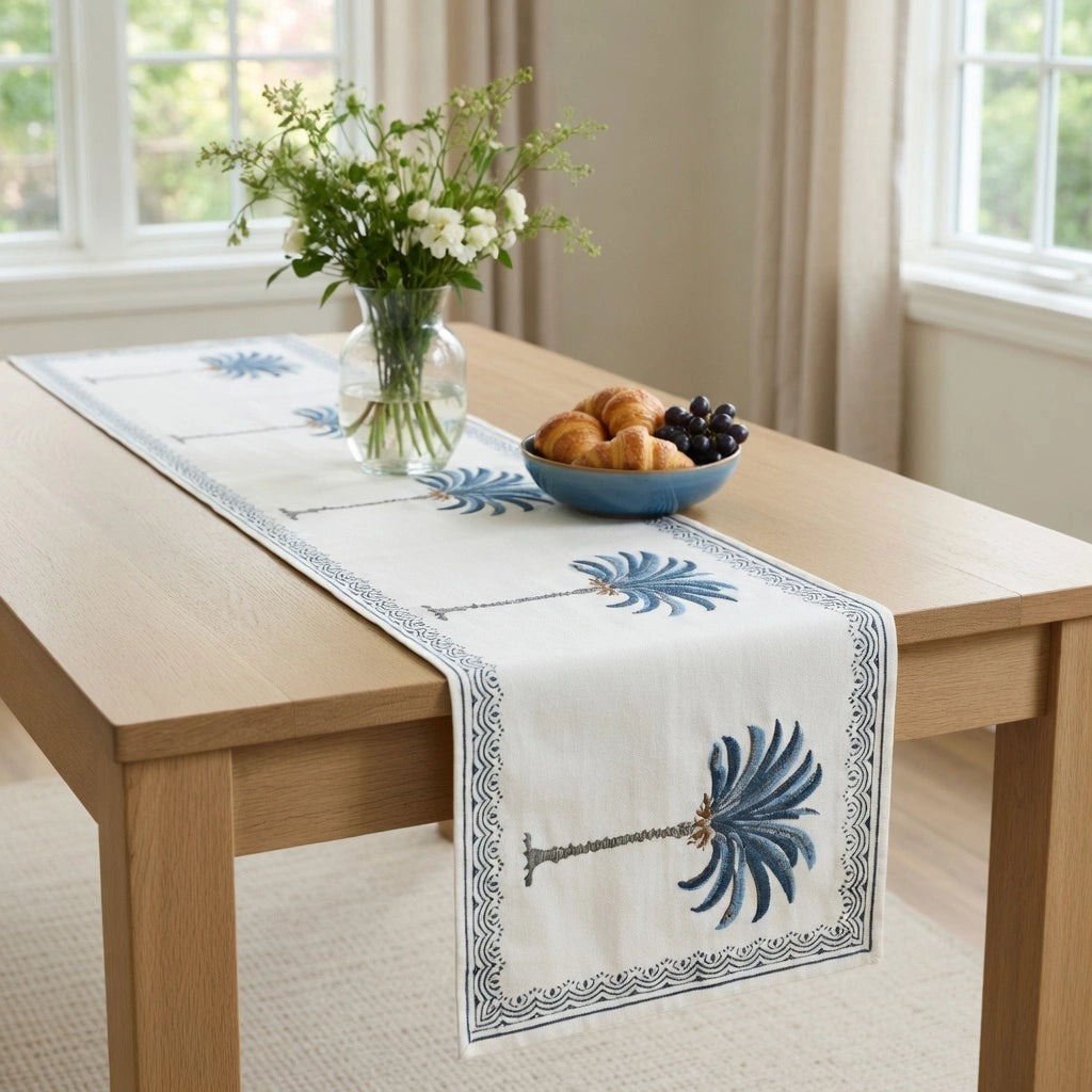 A white table runner with blue palm tree and border patterns laid on a light wood dining table in a bright room. A glass vase with white flowers and a blue bowl of pastries and grapes sit on the table.