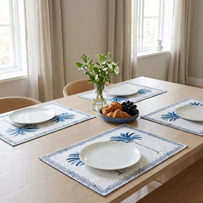 A dining table set with four white placemats featuring blue palm tree and border patterns, styled with white plates, a small vase of flowers, and a bowl of pastries in a bright room with beige curtains.