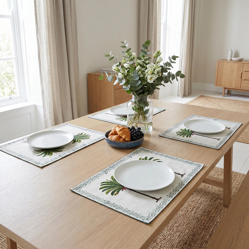 A dining table set with four rectangular placemats featuring a tropical palm leaf pattern in green and blue on a cream background. The table is set with white plates, a glass vase of flowers, and a bowl of pastries, in a bright, modern dining room with beige curtains and wood furniture.