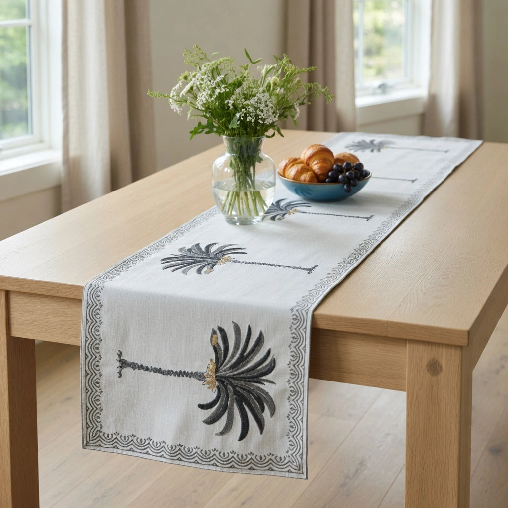 A white cotton table runner with black palm tree prints and a decorative border, styled on a light wood dining table with a vase of flowers and a bowl of fruit.