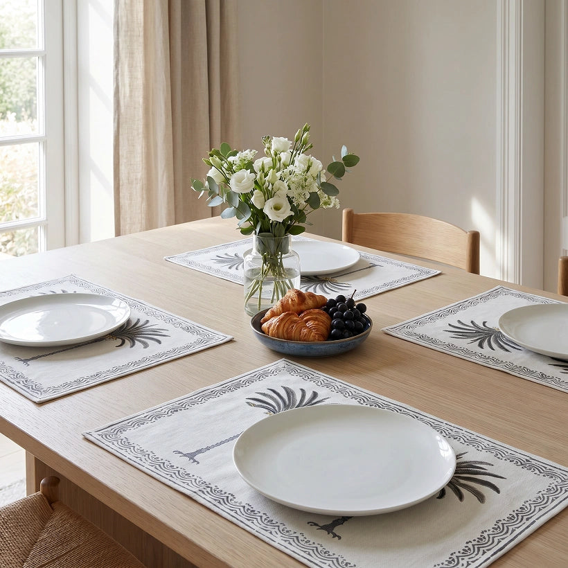 A dining table set with four cream placemats featuring a black palm tree and border pattern, white plates, a vase of white flowers, and a bowl of croissants and grapes in a bright, sunlit room.