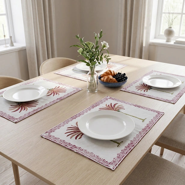 Three rectangular table placemats with a white base and a reddish-brown palm leaf pattern, arranged on a light wood dining table set with white plates, a vase of flowers, and a bowl of fruit.