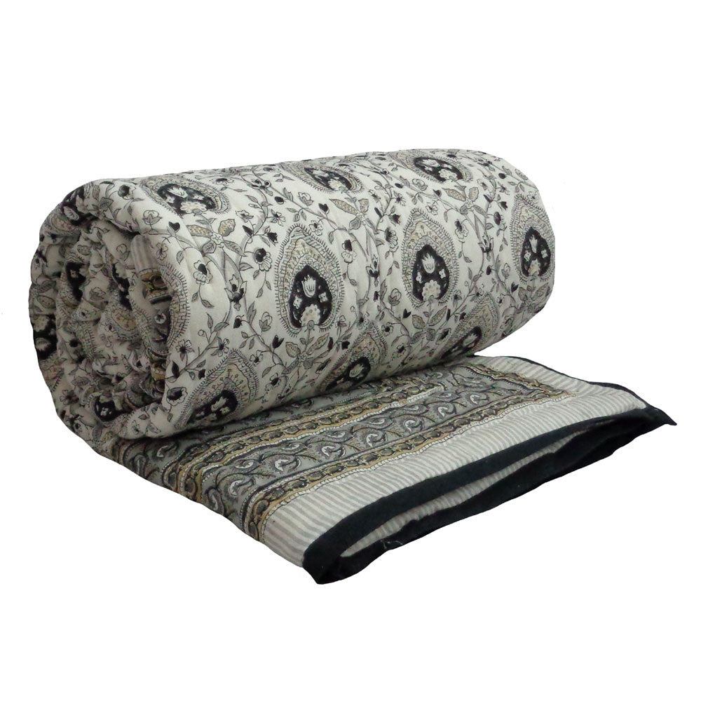 A rolled-up quilted blanket featuring a detailed paisley pattern in black, gray, and beige on a cream background, with a striped border and black piping.