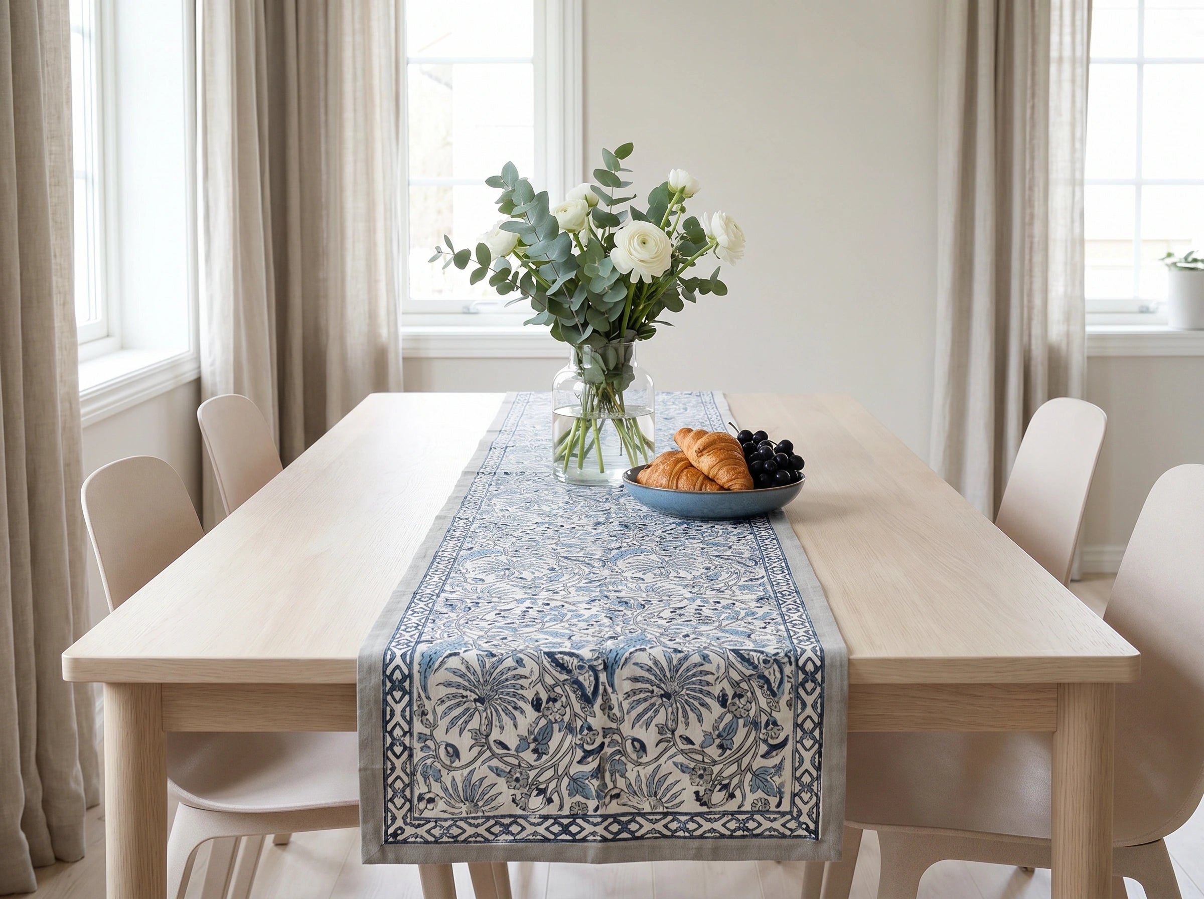 A blue and white patterned table runner with a botanical design is styled on a light wood dining table in a bright, modern room. The table is surrounded by light beige chairs, decorated with a vase of white roses and eucalyptus, and a blue plate with croissants and blackberries.