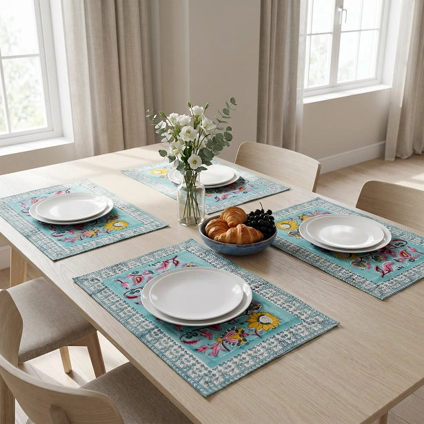 Four rectangular table placemats with a turquoise blue border and colorful floral motifs are arranged around a light wood dining table set with white plates, a bowl of pastries, and a vase of flowers in a bright room.