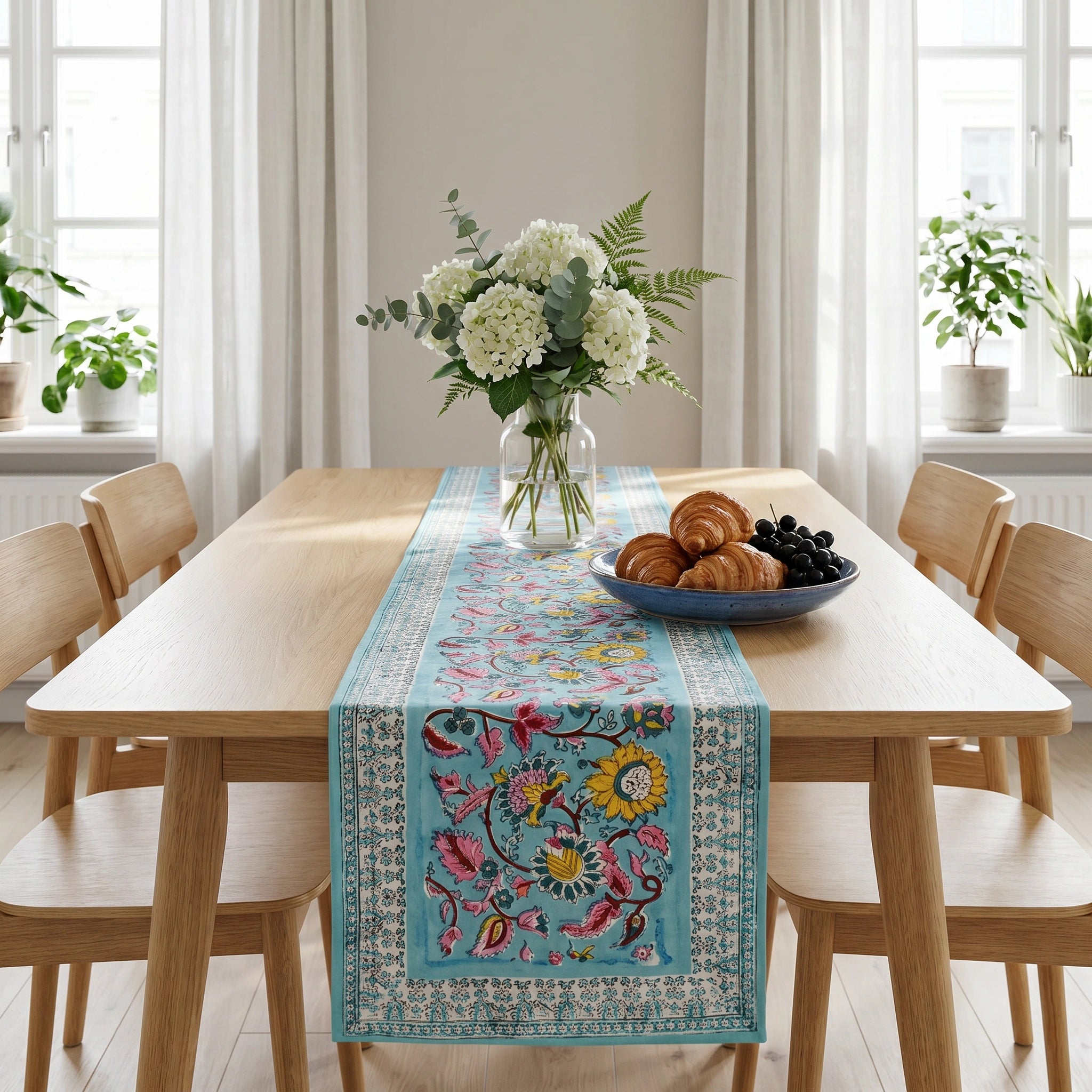 A light blue table runner with a colorful floral and bird pattern, styled on a wooden dining table in a bright, modern room with chairs, a floral centerpiece, and a plate of pastries.