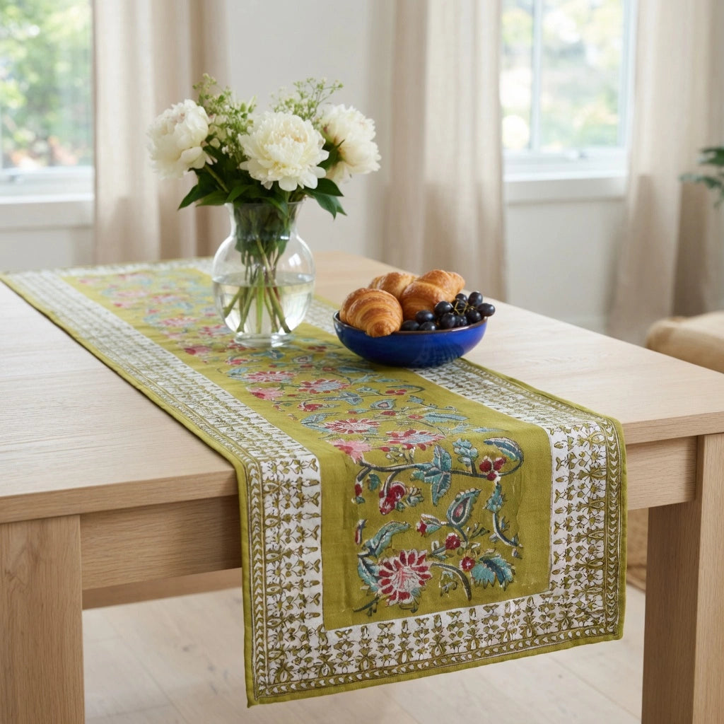 An embroidered table runner in olive green with floral and butterfly motifs, styled on a light wood dining table with a blue bowl of pastries and a vase of white flowers.