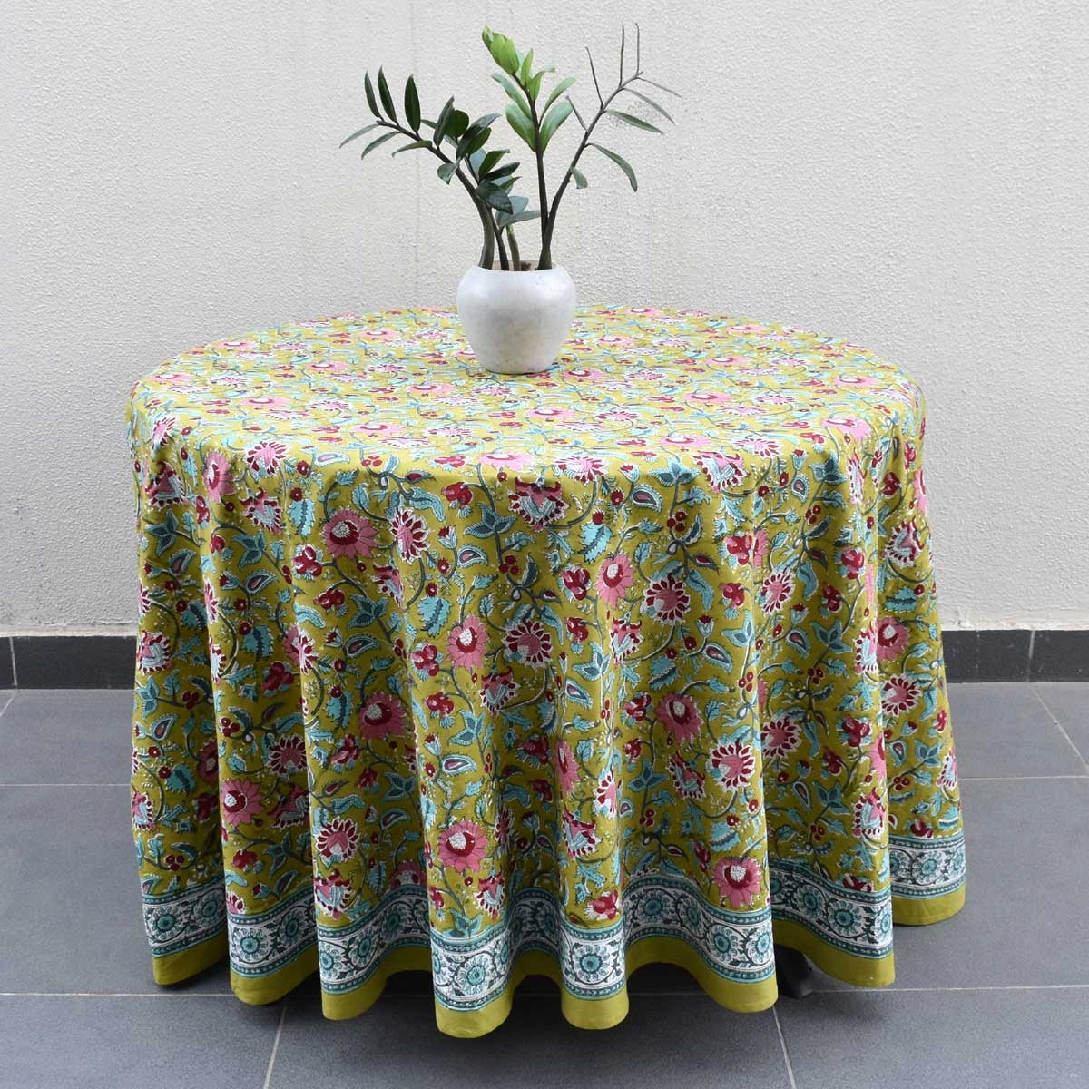 A round table covered with a vibrant green tablecloth featuring a colorful floral and paisley pattern, styled with a white vase containing green leaves, set against a neutral textured wall and gray tiled floor.