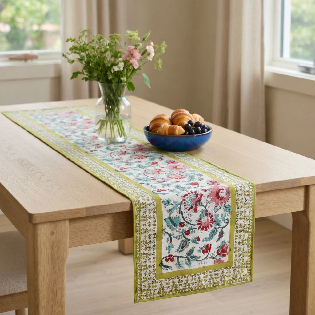 A floral table runner with green borders is styled on a light wood dining table, alongside a blue bowl of pastries and a vase of flowers in a bright room.