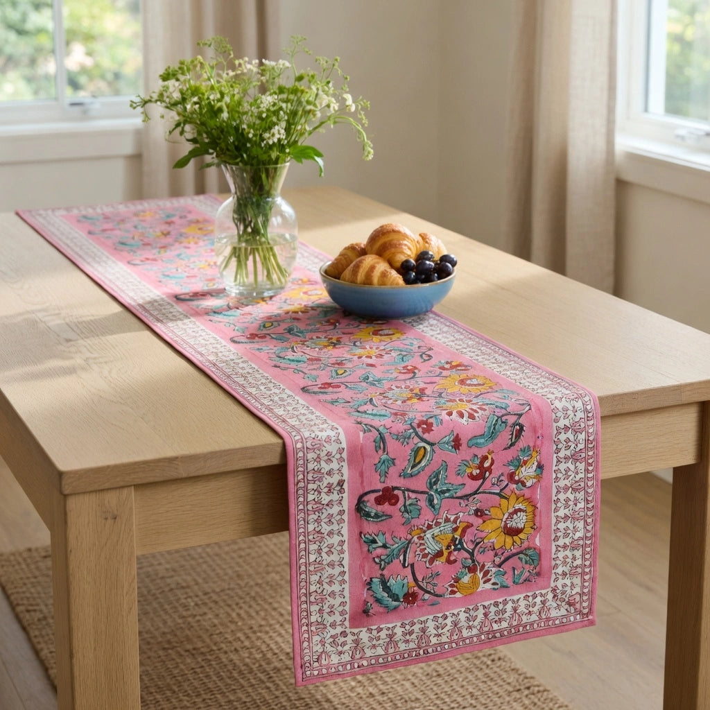 A pink embroidered table runner with a floral and bird pattern, draped over a light wood dining table. The runner features white borders and colorful floral embroidery.