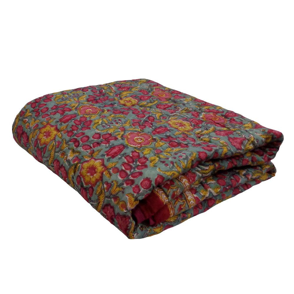 A folded quilted blanket featuring a vibrant floral pattern with pink, red, and yellow flowers on a muted blue-gray background, set against a plain white studio backdrop.