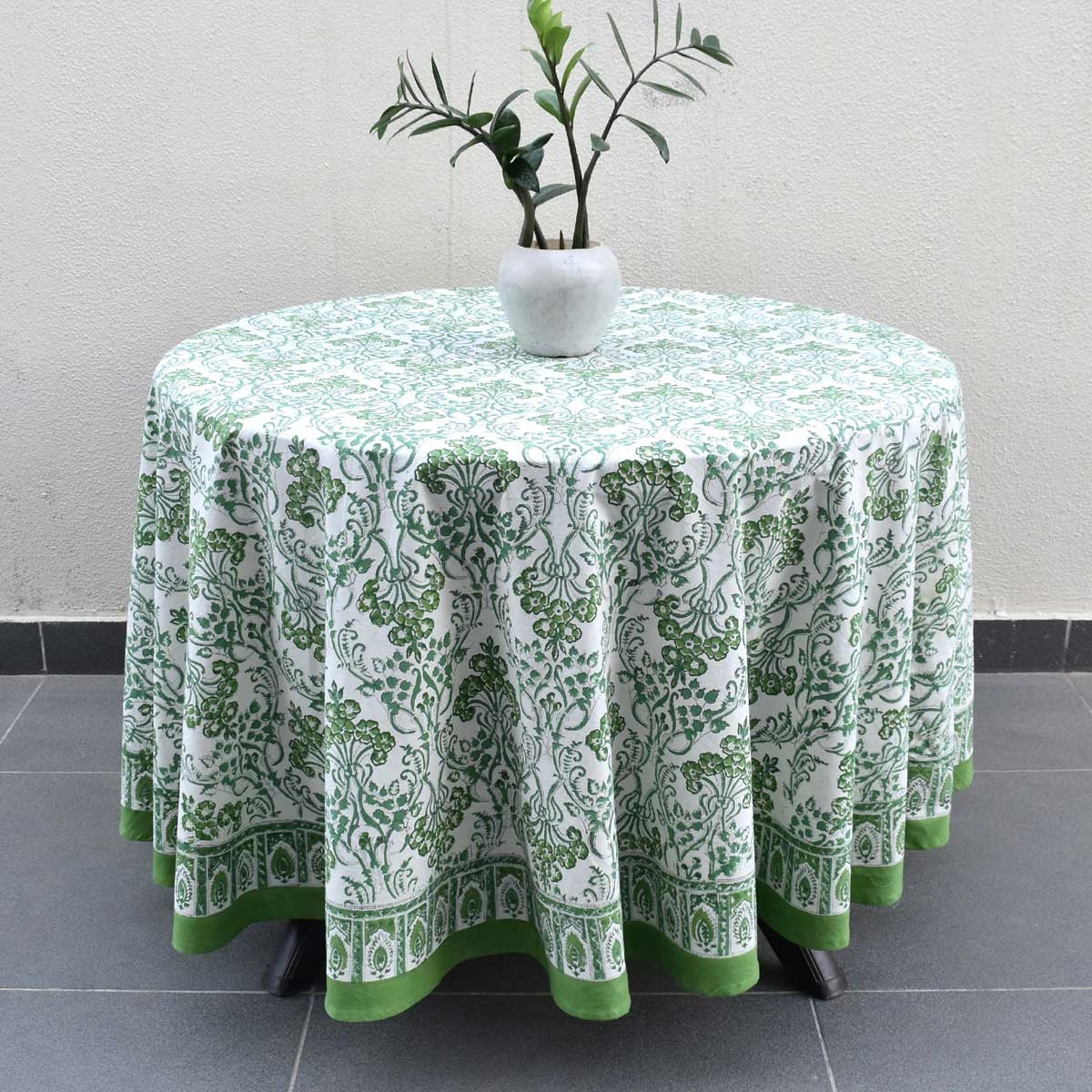 A round table covered with a white tablecloth featuring a green botanical damask pattern and a solid green border, styled with a small white vase of green leaves on top.