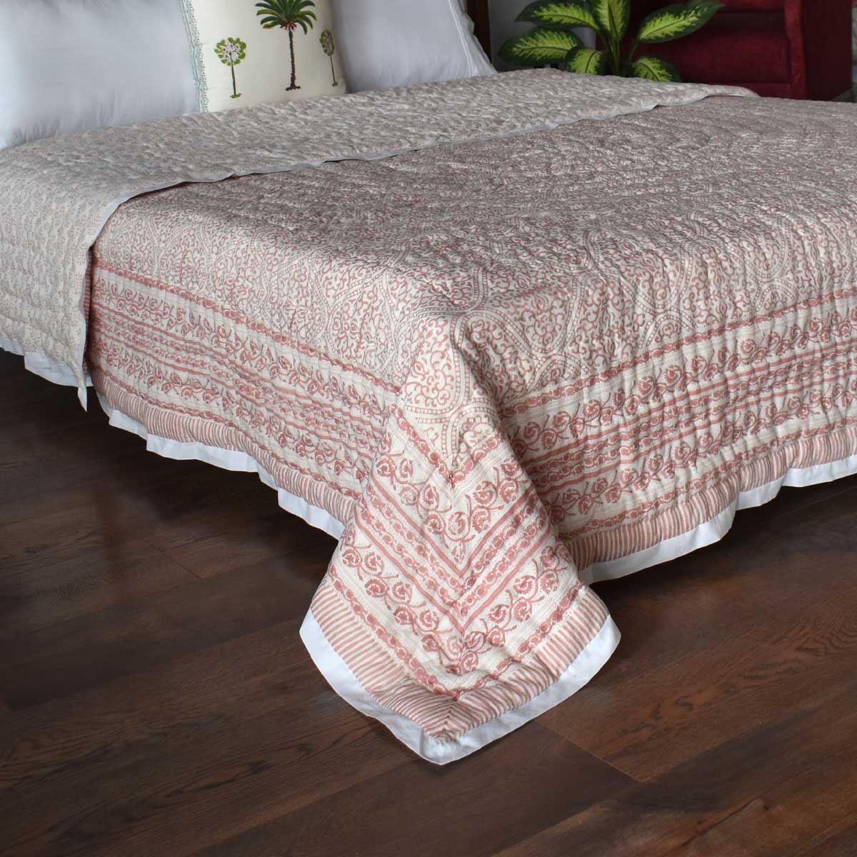 A quilted bedspread with a pink and cream bohemian pattern, draped over a bed on a dark wood floor with white pillows visible in the background.