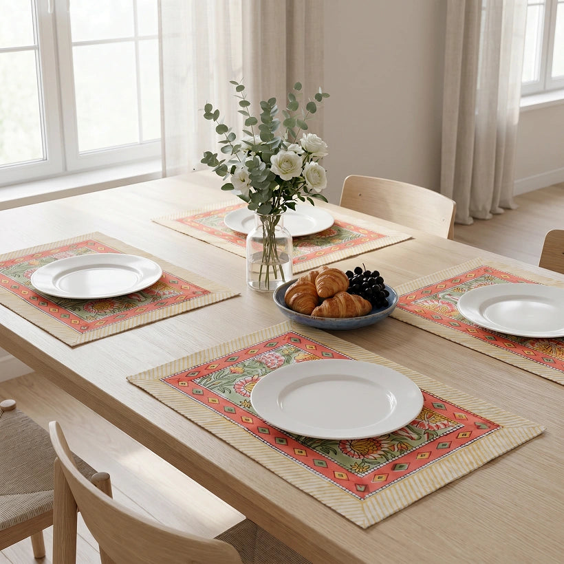A dining table set with four matching rectangular placemats featuring a colorful floral and geometric pattern, white plates, a vase of flowers, and a bowl of pastries in a bright, modern dining room.