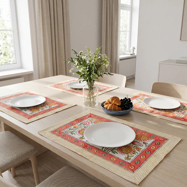 Three rectangular woven placemats with a colorful floral and geometric border pattern are arranged on a light wood dining table. Each placemat is styled with a white plate, and the table is further decorated with a glass vase of green stems and a small bowl of pastries and grapes.