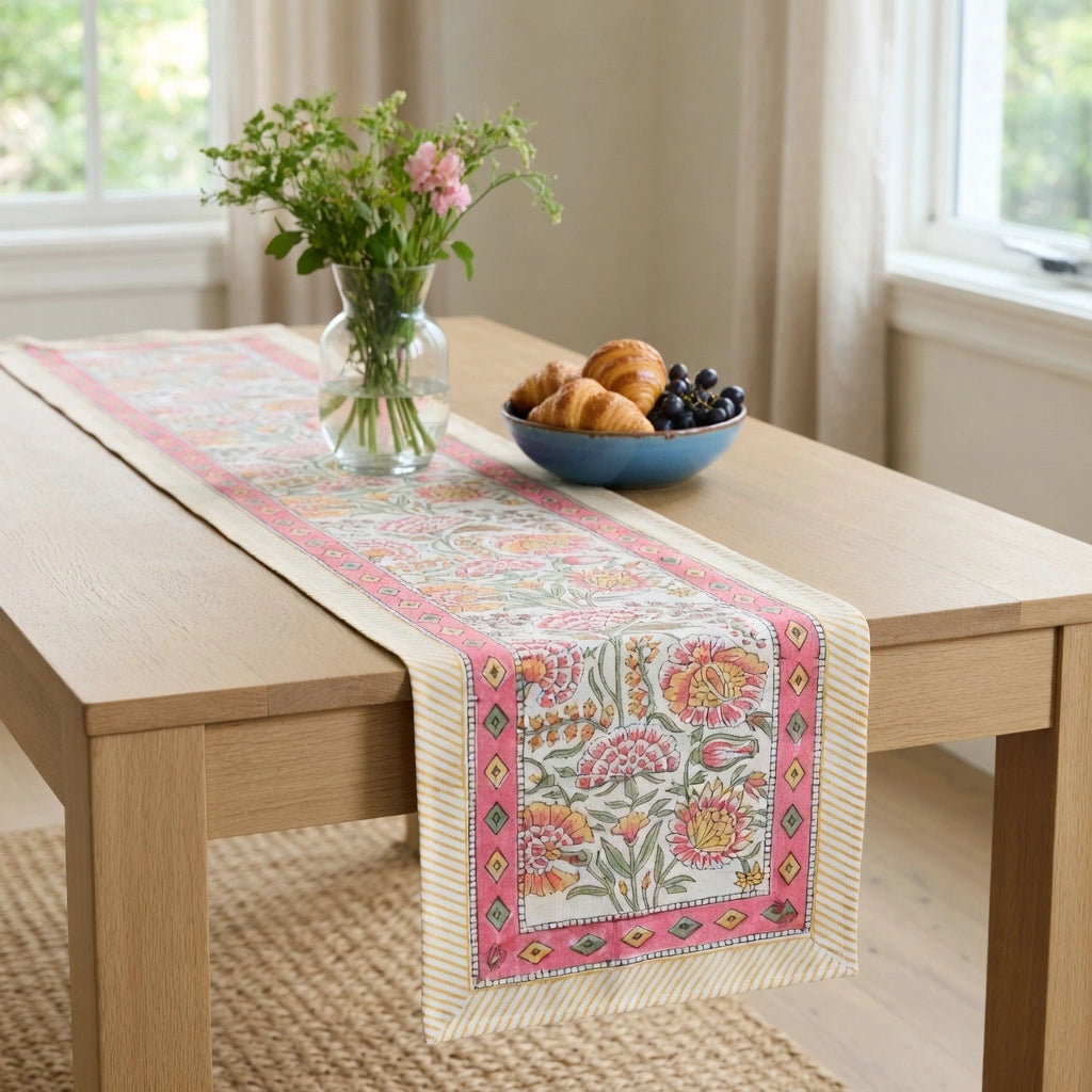 A decorative table runner with a pink and cream floral pattern is styled on a light wood dining table, flanked by a glass vase of flowers and a blue bowl of pastries in a bright room.