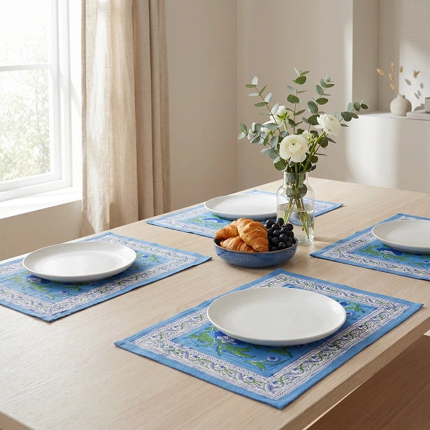 Four blue patterned placemats arranged on a light wood dining table, each styled with a white plate, with a vase of flowers and a bowl of pastries in the center of the scene.
