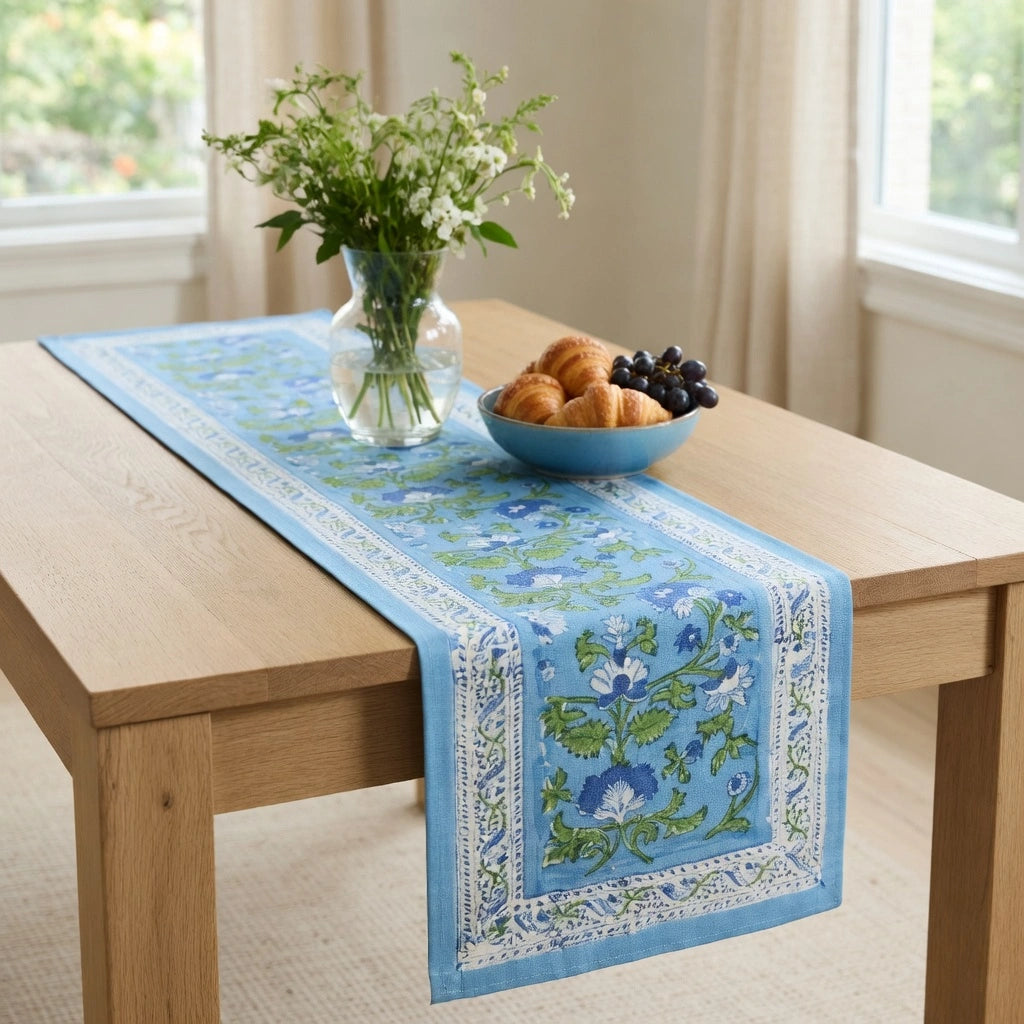 A light blue table runner with a floral pattern and white border, styled on a light wood dining table with a blue bowl of pastries and a vase of flowers.
