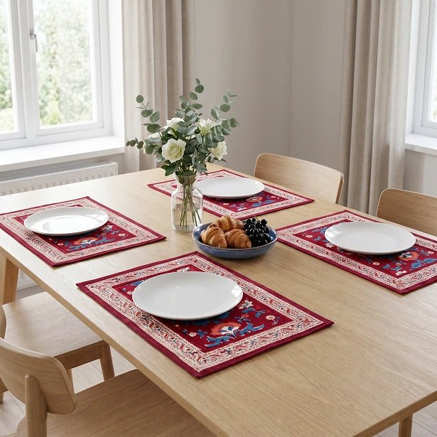 Four rectangular table placemats with red borders and blue, yellow, and white traditional patterns are arranged on a light wood dining table set with white plates, a vase of flowers, and a bowl of pastries in a bright room.