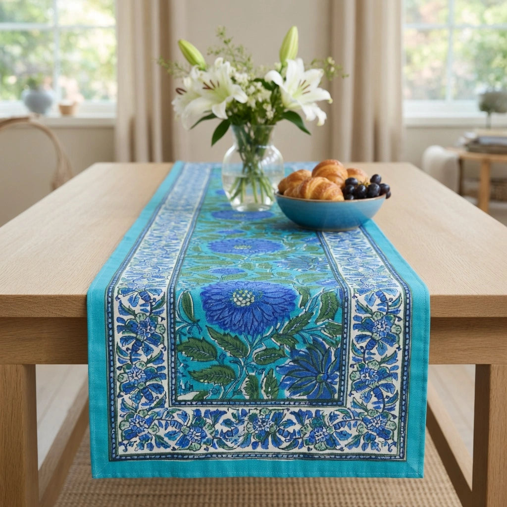 A blue and turquoise floral table runner with white border details is styled on a light wood dining table, flanked by a blue bowl of pastries and a vase of white lilies in a bright, sunlit room.